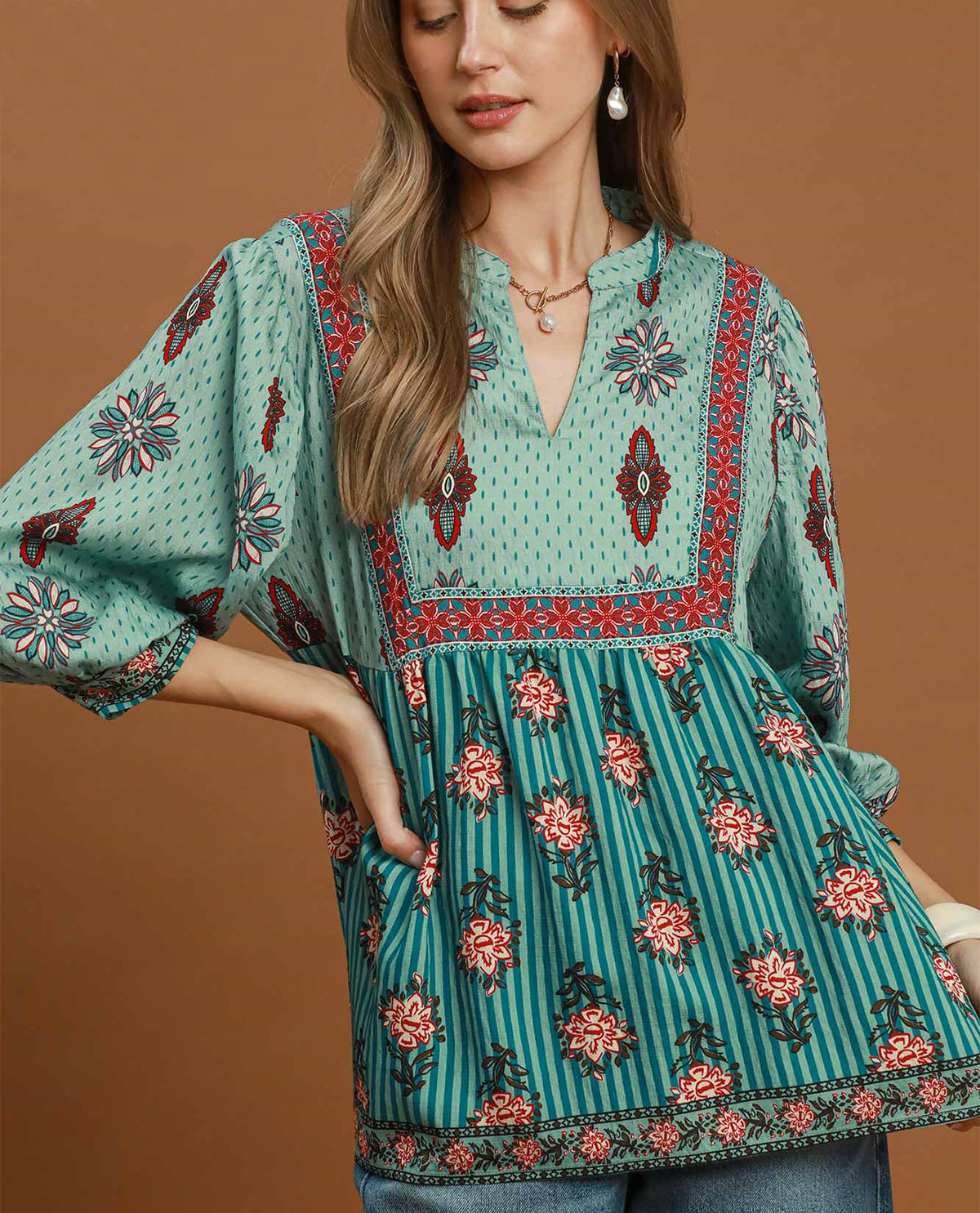 A woman wearing a bohemian-style tunic top with a split design: a light mint green dotted yoke with red and navy floral embroidery, and a teal-green striped skirt with pink and red floral prints. The background is a solid warm brown studio backdrop.