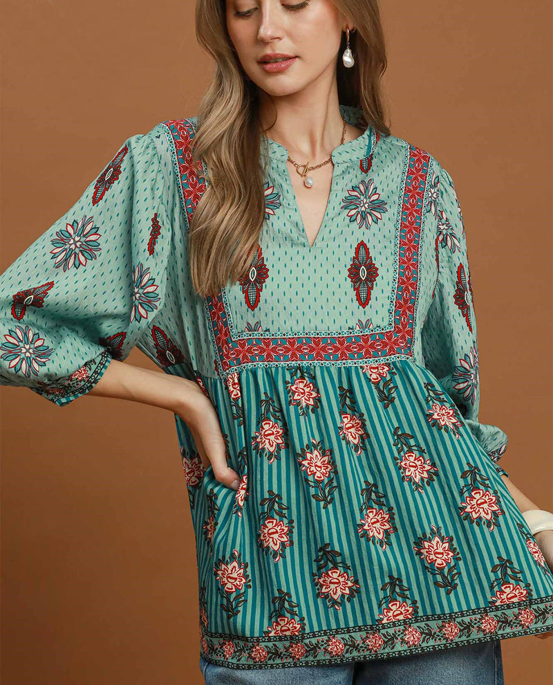 A woman wearing a bohemian-style tunic top with a split design: a light mint green dotted yoke with red and navy floral embroidery, and a teal-green striped skirt with pink and red floral prints. The background is a solid warm brown studio backdrop.