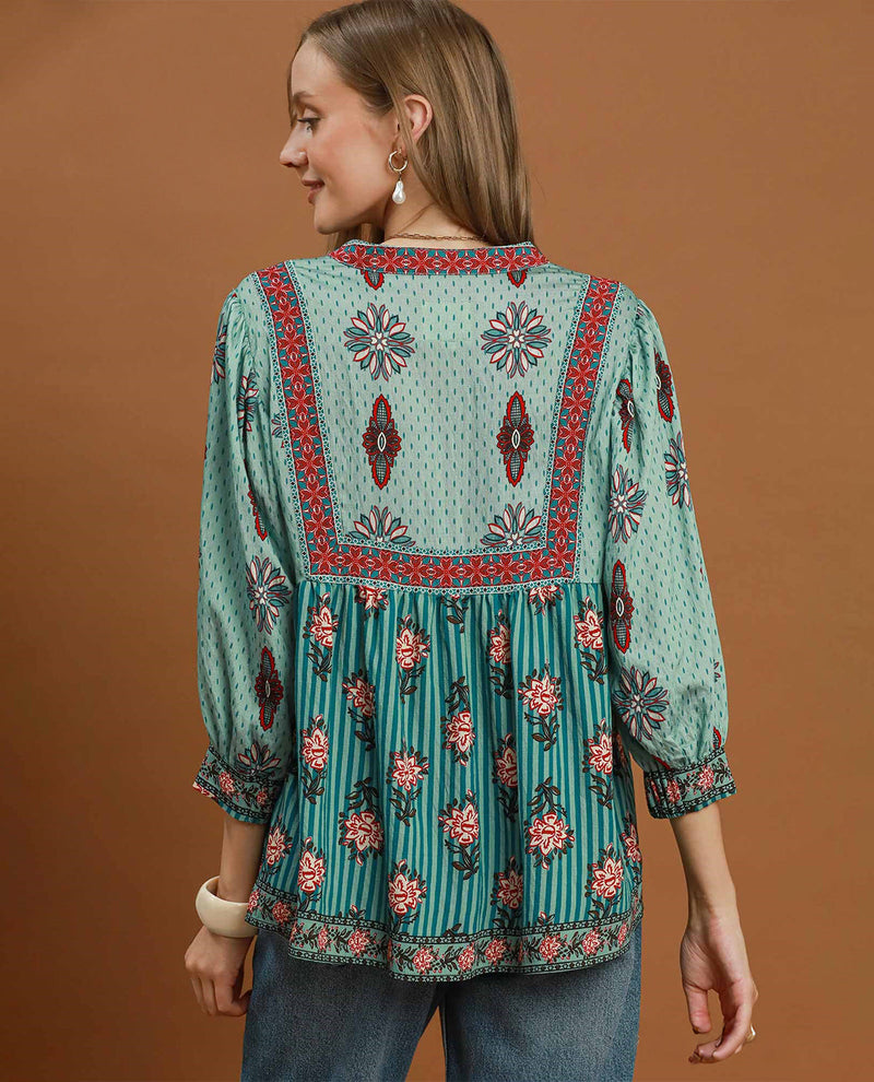 A woman shown from the back wearing a teal green bohemian-style top with red and black geometric borders and pink floral patterns, paired with blue jeans, against a solid brown studio background.