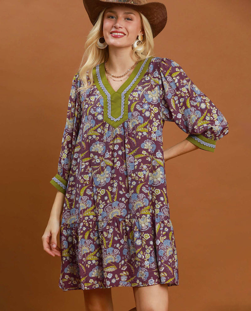 A woman wearing a bohemian-style tiered dress with a purple, blue, and yellow floral pattern, olive green trim, and puff sleeves, paired with a brown cowboy hat, set against a solid brown studio background.