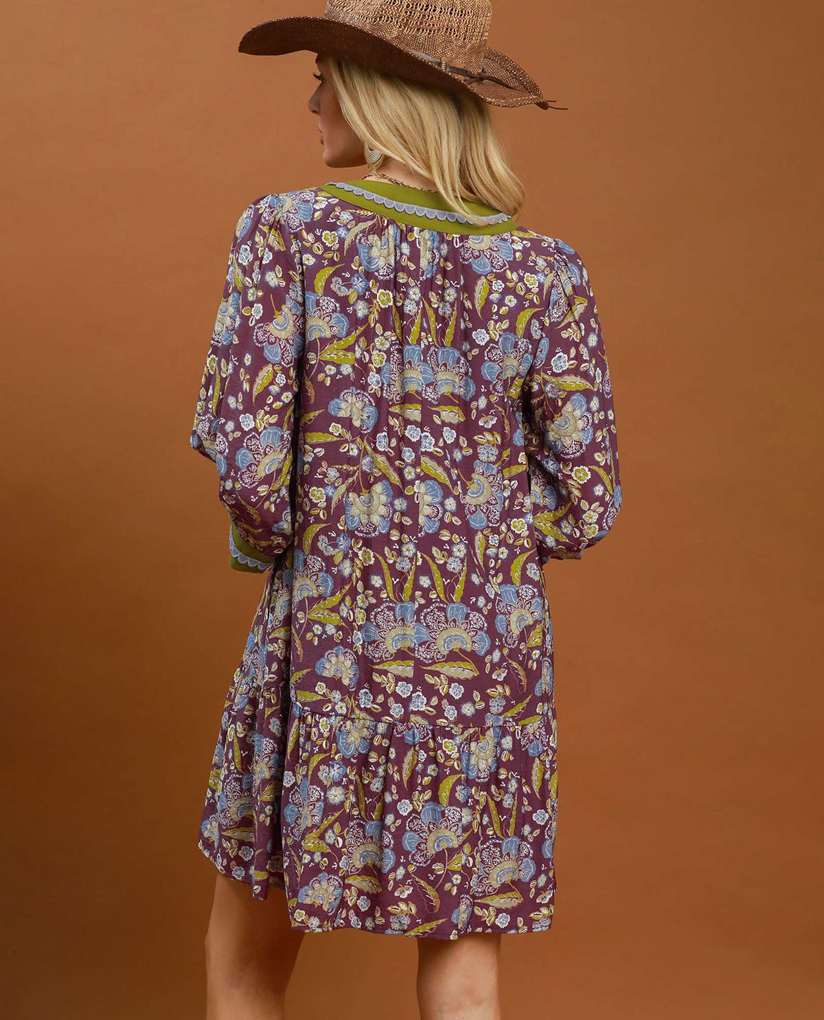 A woman wearing a bohemian-style tiered dress with a purple, yellow, and blue floral pattern, shown from the back against a solid brown studio background.