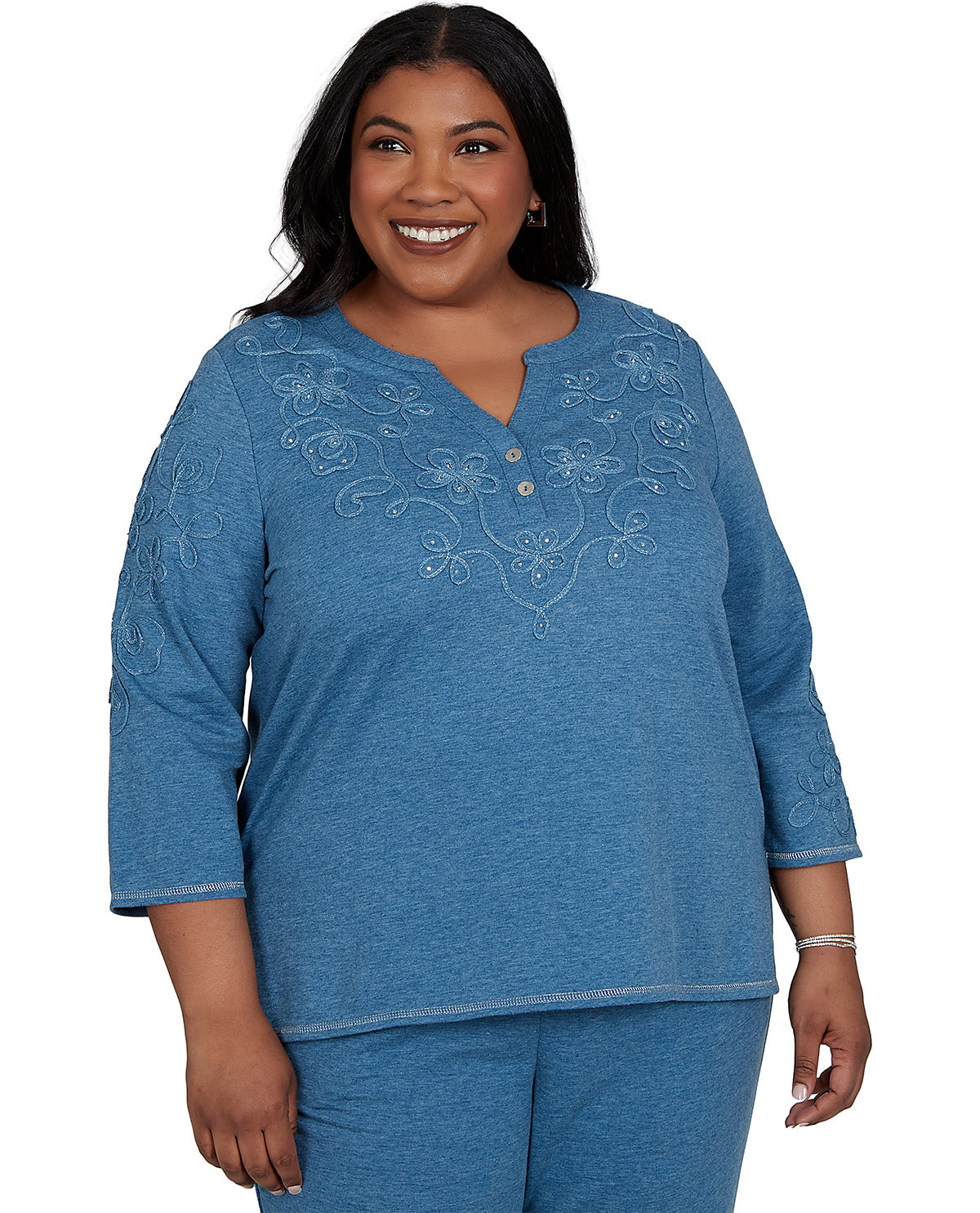 A smiling woman wearing a heathered blue three-quarter sleeve top with white floral embroidery and a buttoned split neckline, paired with matching blue pants, against a plain white background.