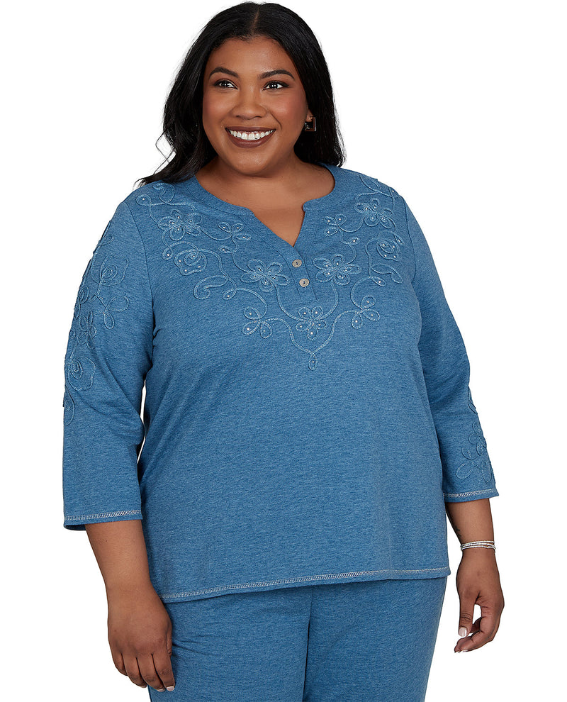A smiling woman wearing a heathered blue three-quarter sleeve top with white floral embroidery and a buttoned split neckline, paired with matching blue pants, against a plain white background.