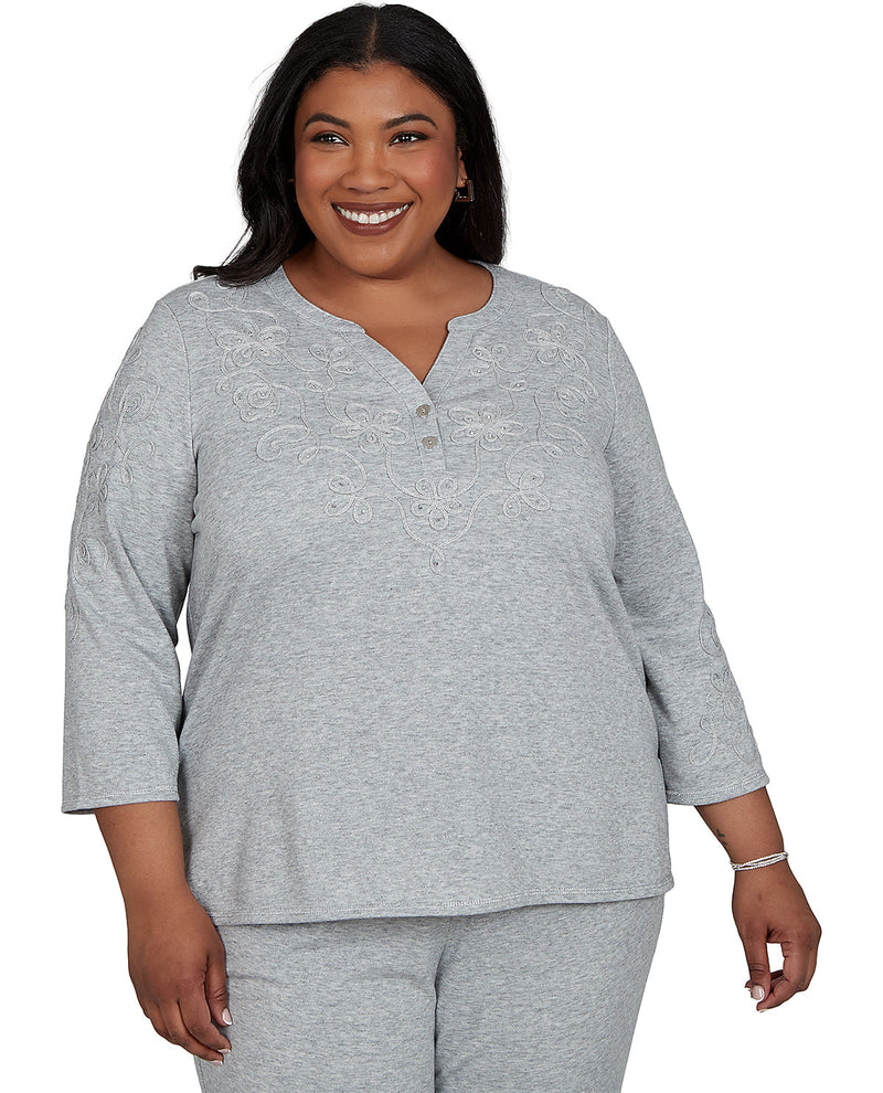 A smiling woman modeling a light gray heather three-piece loungewear set, featuring a henley top with embroidered detailing on the chest, matching sweatpants, and a matching belt, shot against a plain white background.