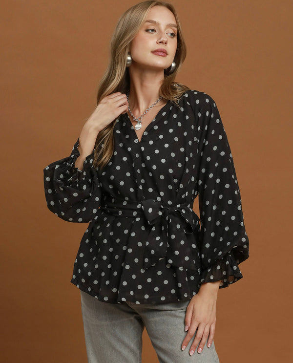 A woman modeling a black long-sleeve blouse with a white polka dot pattern, featuring a V-neckline, tie waist, and ruffled cuffs, set against a solid brown studio background.