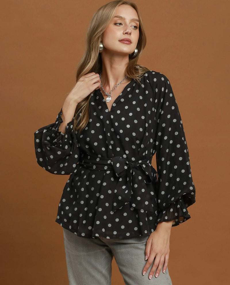 A woman modeling a black long-sleeve blouse with a white polka dot pattern, featuring a V-neckline, tie waist, and ruffled cuffs, set against a solid brown studio background.