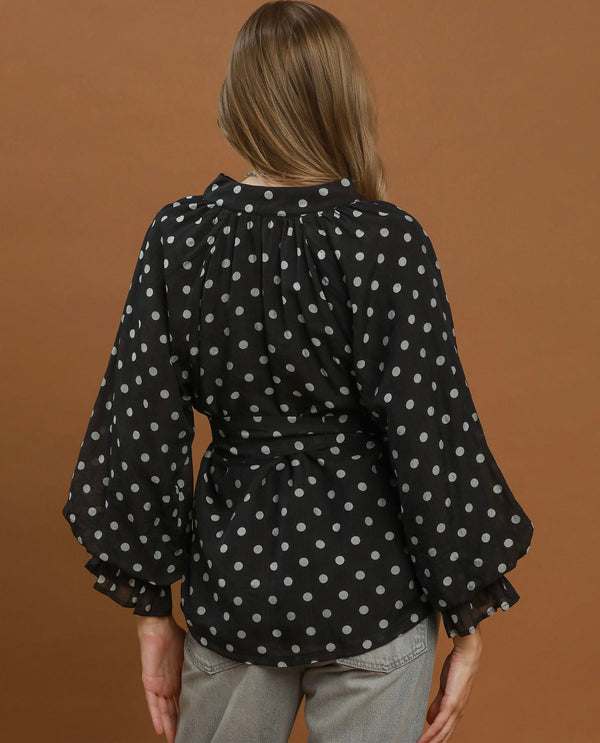 A woman viewed from the back wearing a black long-sleeve blouse with white polka dots, gathered at the shoulders and waist, paired with light gray pants, against a solid brown background.