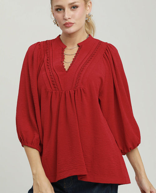 A woman wearing a red long-sleeve blouse with a gathered peplum waist, scalloped keyhole neckline, and puff sleeves, paired with dark blue jeans against a light gray studio background.