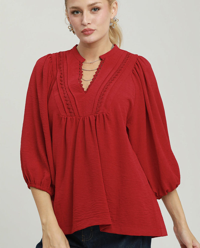 A woman wearing a red long-sleeve blouse with a gathered peplum waist, scalloped keyhole neckline, and puff sleeves, paired with dark blue jeans against a light gray studio background.