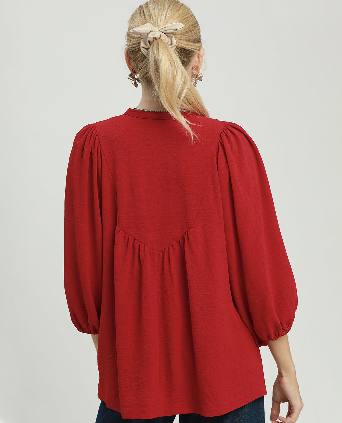 A woman viewed from the back wearing a red long-sleeve blouse with voluminous puff sleeves and gathered detailing at the waist, paired with dark blue jeans against a light gray studio background.