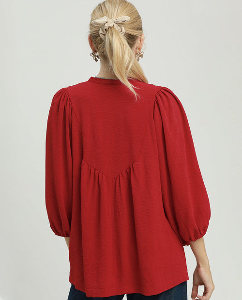 A woman viewed from the back wearing a red long-sleeve blouse with voluminous puff sleeves and gathered detailing at the waist, paired with dark blue jeans against a light gray studio background.