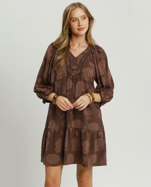 A woman modeling a brown tiered mini dress with a tonal abstract pattern, featuring a V-neckline and voluminous elbow-length sleeves, set against a plain light gray studio background.