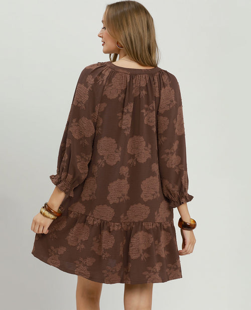 A woman shown from the back wearing a brown long-sleeve dress with a darker brown floral pattern, puff sleeves, and a ruffled hem, set against a plain light gray studio background.