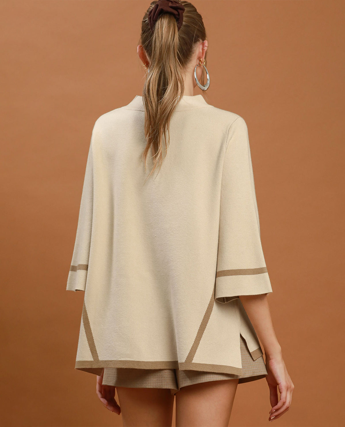 A woman wearing a cream-colored knit poncho-style top with wide three-quarter sleeves and contrasting beige stripes near the cuffs, posed against a solid warm brown background.