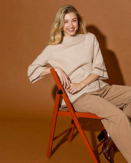A woman sits on a rust-orange folding chair against a warm brown studio backdrop, wearing a beige mock-neck top with wide flared sleeves and matching beige trousers.