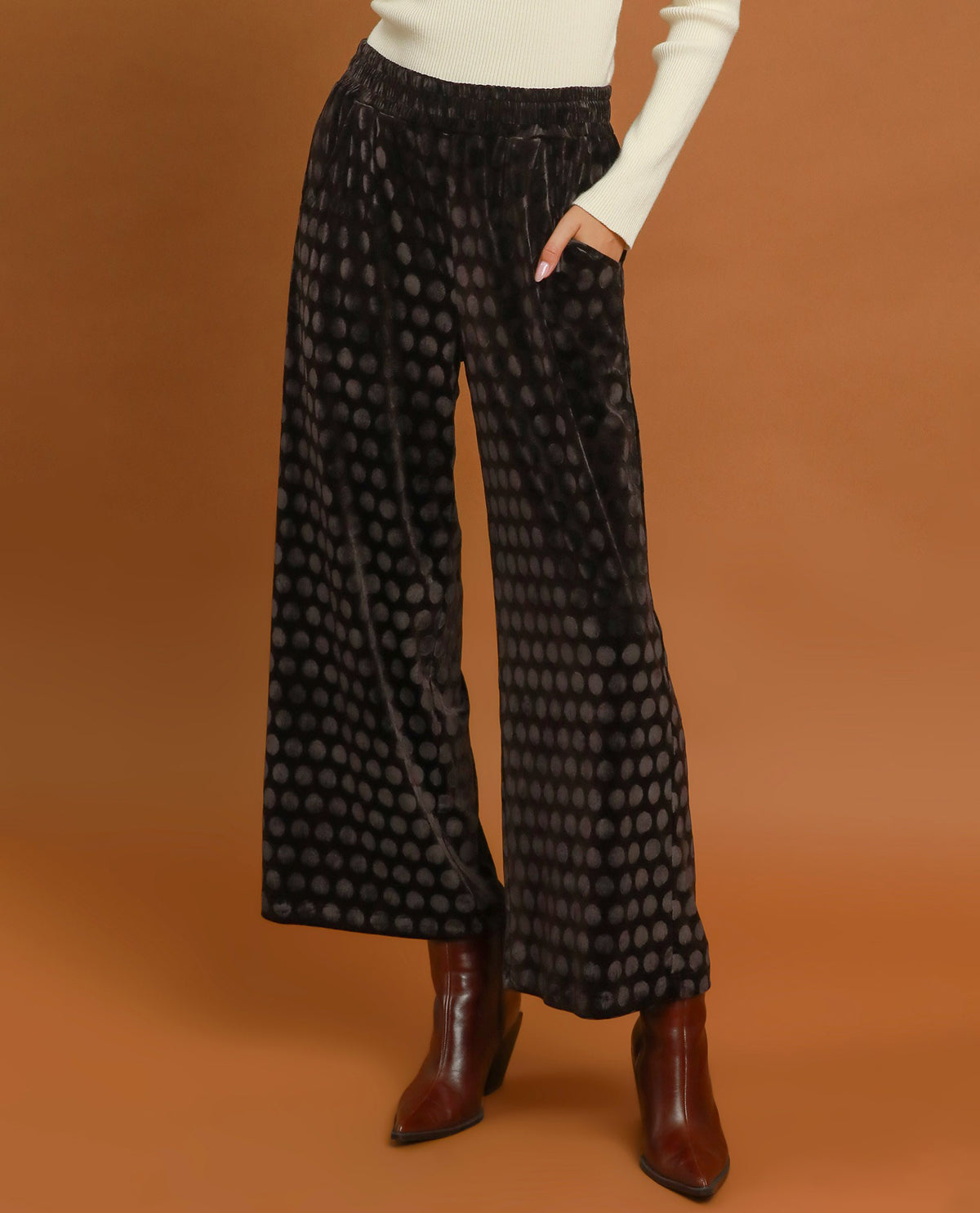 Black wide-leg velvet pants with a dark gray polka dot pattern, styled with a cream long-sleeve top and brown leather ankle boots against a solid warm brown background.