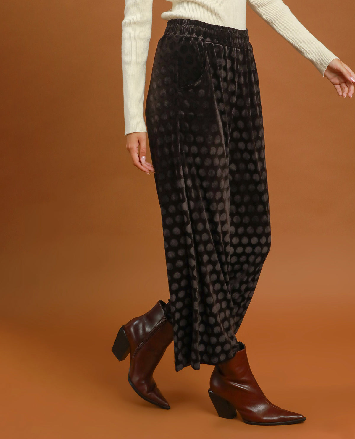 A person wearing loose black velvet pants with a subtle circular pattern, styled with a cream long-sleeve top and brown ankle boots against a solid brown studio background.