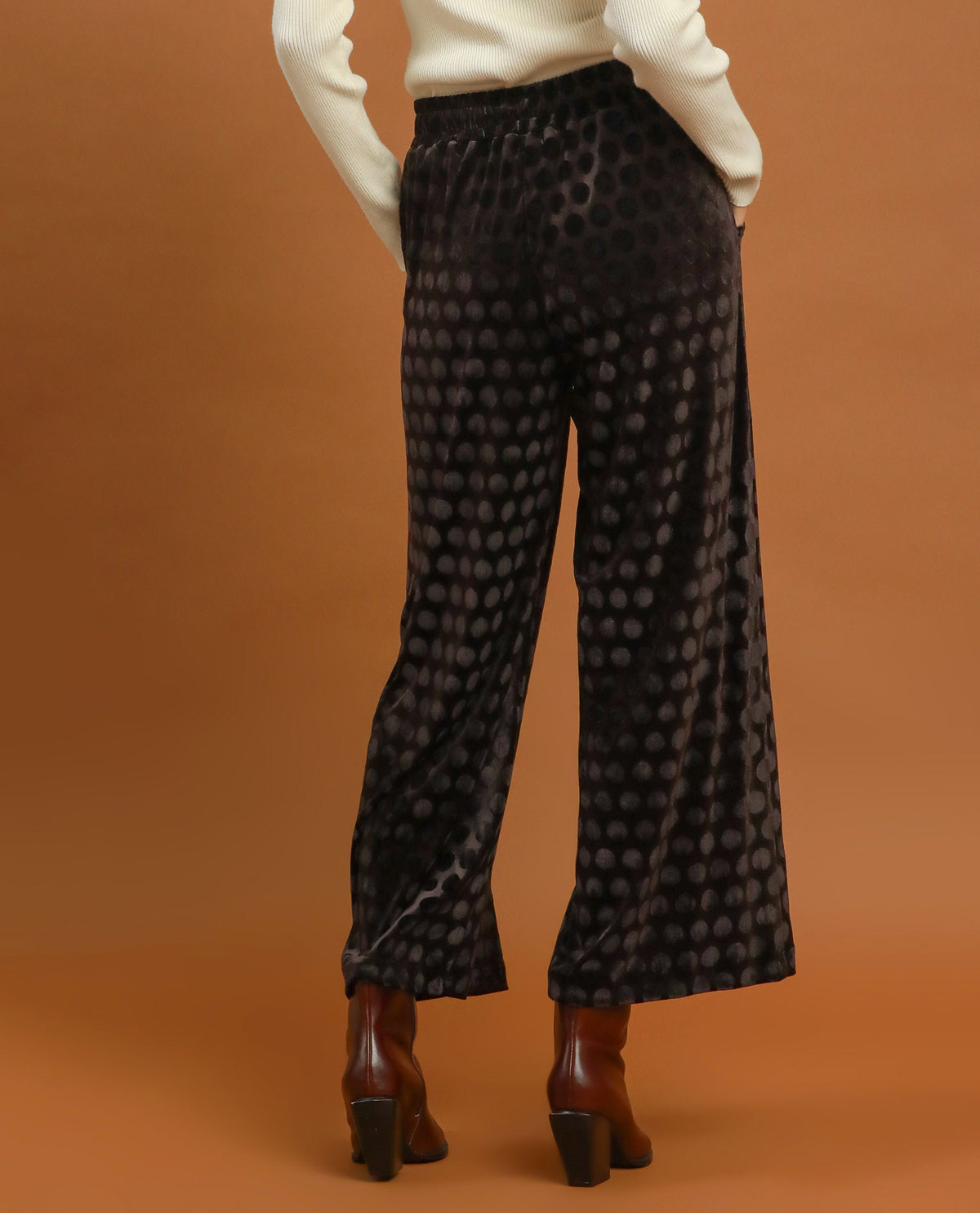 Black wide-leg trousers with a dark gray polka dot pattern, shown on a model from the back against a solid brown studio background.