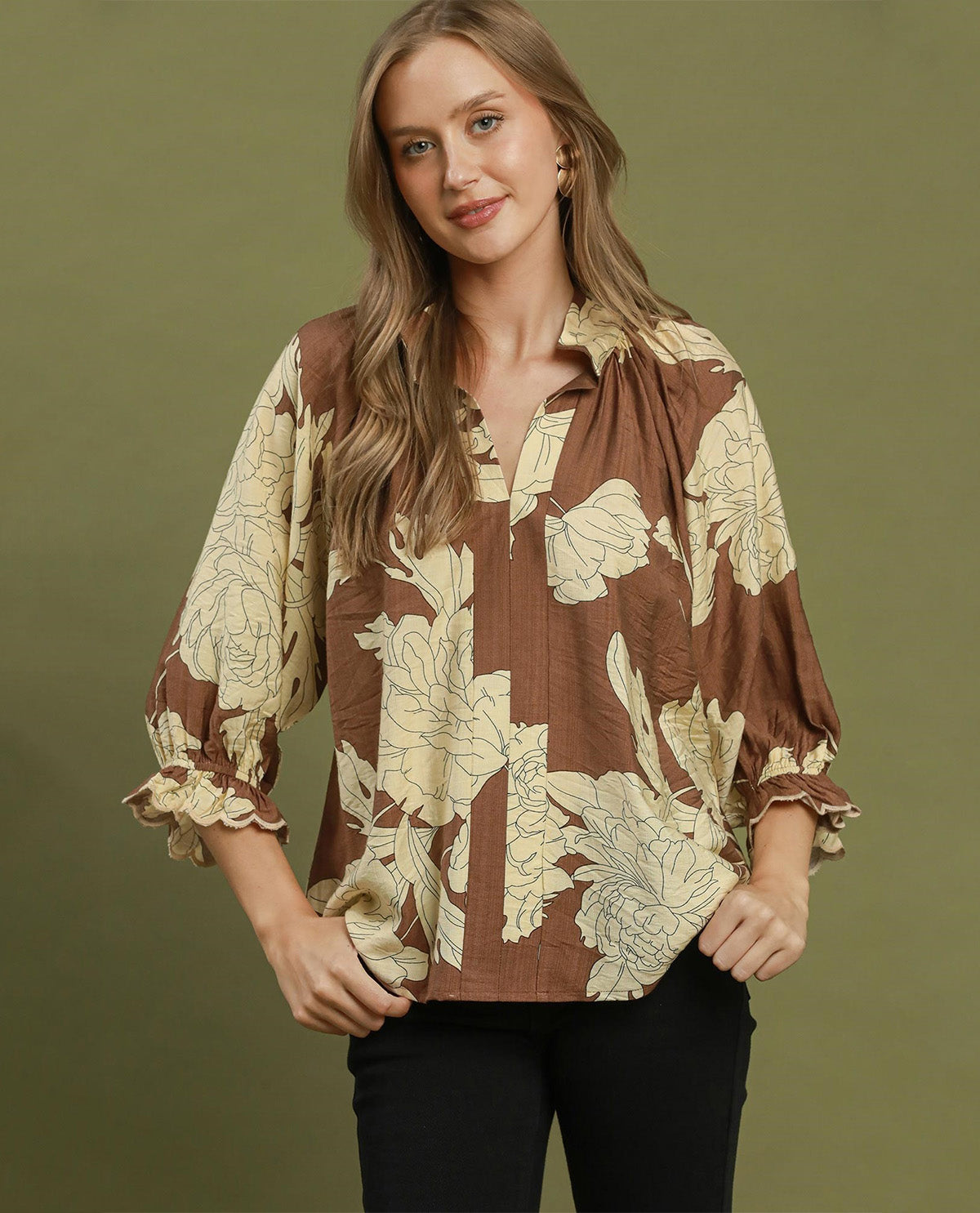 A woman modeling a brown and beige floral print blouse with ruffled sleeves, paired with black pants, against a solid olive green background.