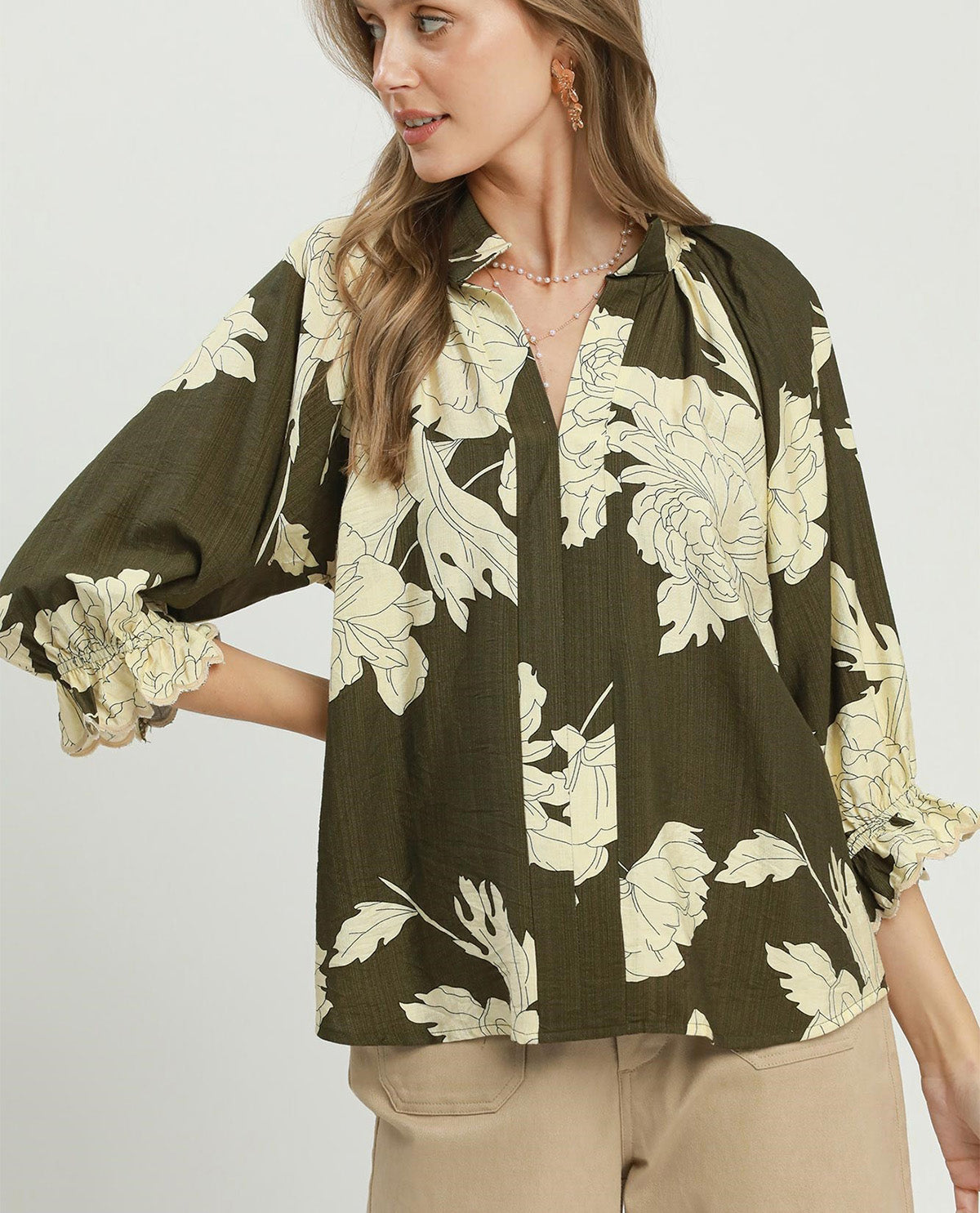 A woman wearing a dark olive green blouse with a large cream floral print, paired with beige pants, against a plain light gray background.