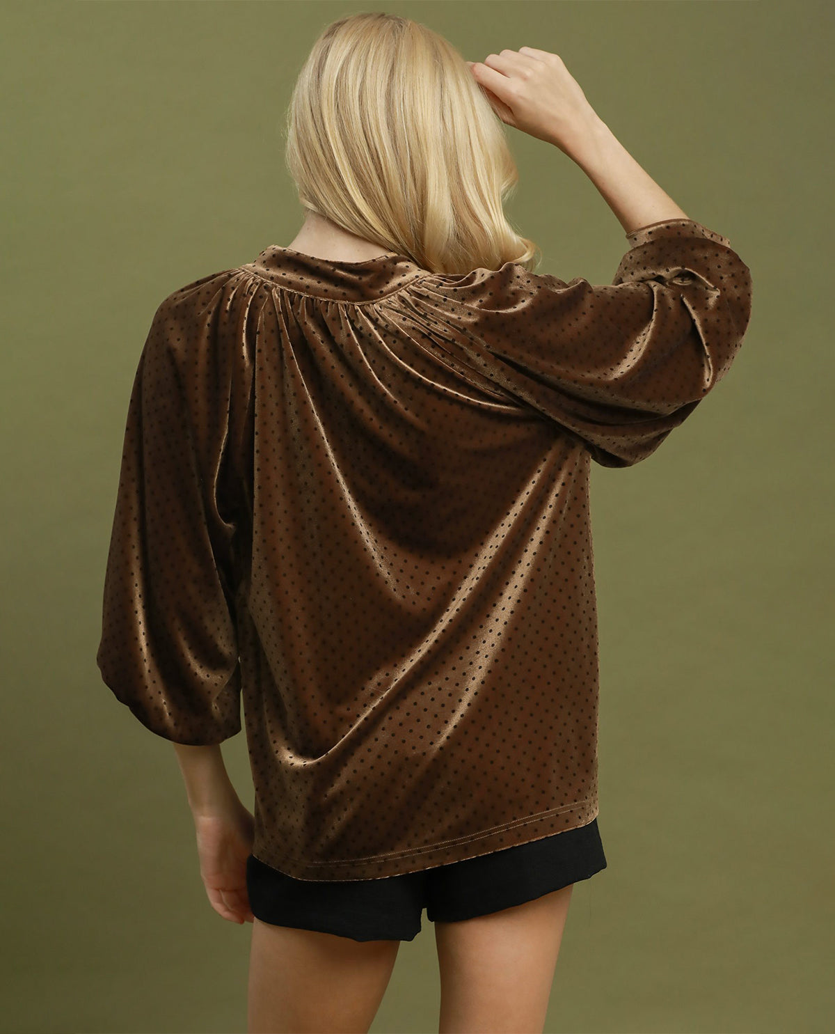A woman shown from the back wearing a brown velvet top with a subtle polka dot pattern and black shorts, set against a solid olive green background.