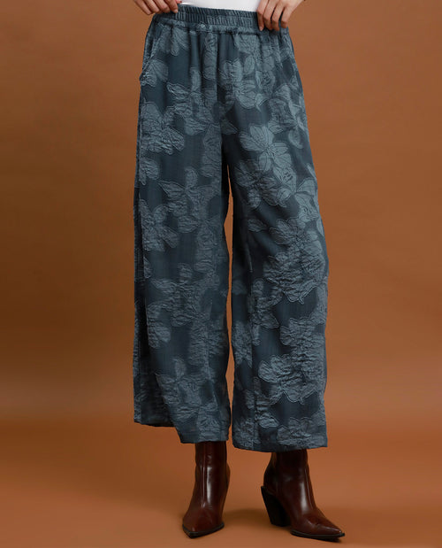 A person wearing wide-leg cropped pants in a muted blue-gray floral jacquard pattern, paired with dark brown ankle boots, against a solid warm brown studio background.