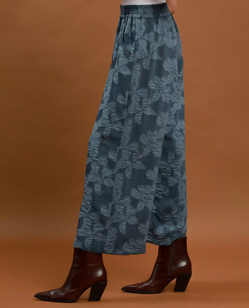 A person shown from the waist down wearing wide-leg cropped pants in a muted blue-gray floral jacquard pattern, paired with dark brown heeled ankle boots, against a solid warm brown studio background.