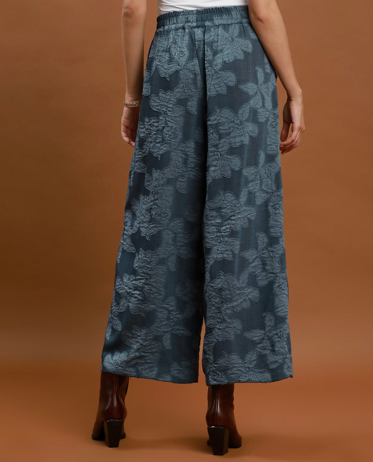 A person shown from the back wearing wide-leg cropped pants in a muted blue-gray floral jacquard fabric, paired with brown heeled ankle boots against a solid warm brown studio backdrop.