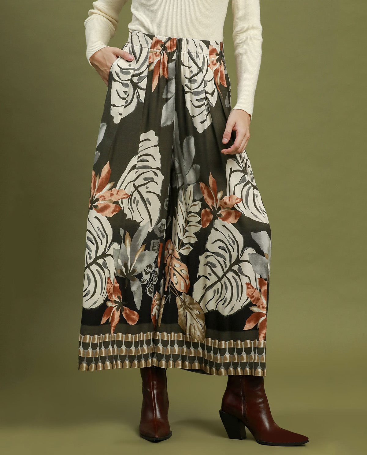A model wearing high-waisted, wide-leg pleated maxi pants with a botanical monstera leaf and orange floral print on a black background, styled with a cream ribbed sweater and dark brown leather ankle boots against a solid olive green backdrop.