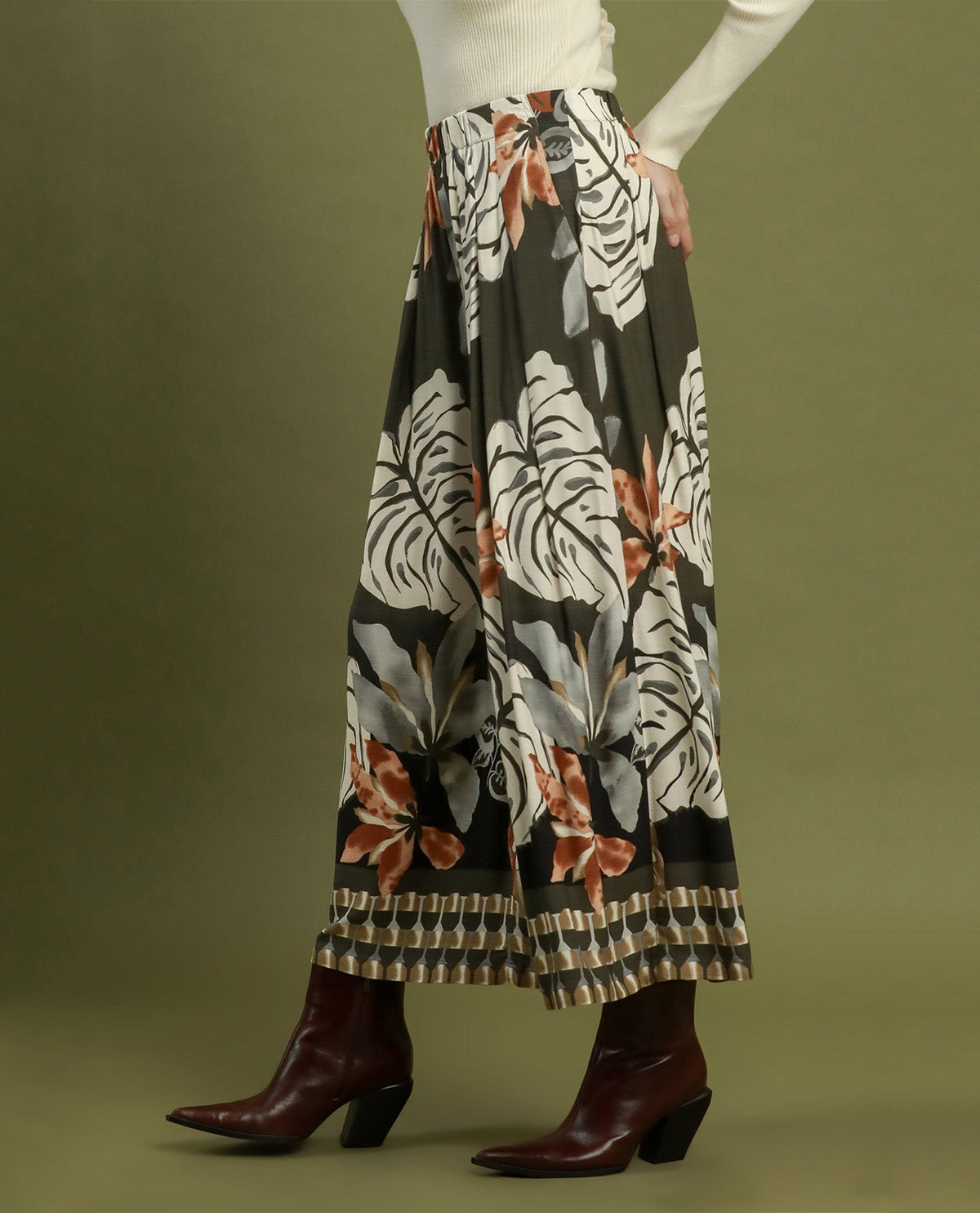 A model is shown from the waist down wearing a wide-leg, pleated maxi skirt with a tropical floral print in white, gray, and rust orange on a black background, paired with dark brown ankle boots against a solid olive green studio backdrop.