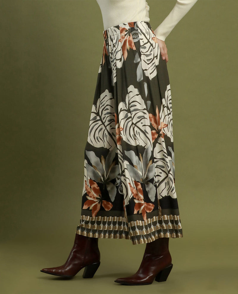A model is shown from the waist down wearing a wide-leg, pleated maxi skirt with a tropical floral print in white, gray, and rust orange on a black background, paired with dark brown ankle boots against a solid olive green studio backdrop.