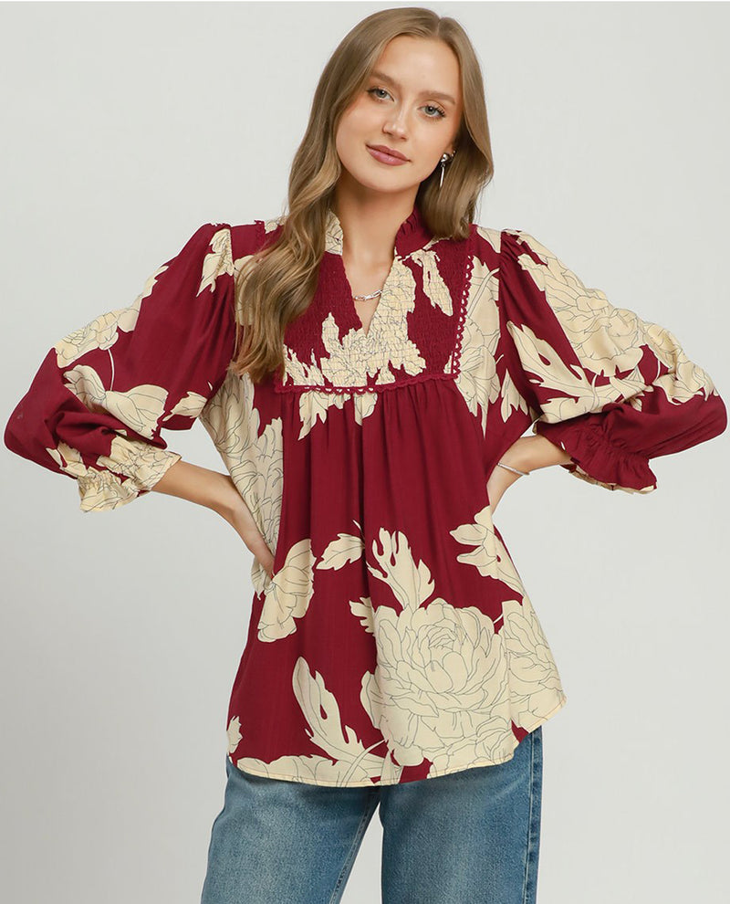 A woman modeling a long-sleeve blouse with a deep V-neck, gathered waist, and voluminous sleeves. The blouse features a deep burgundy base with a cream floral pattern.