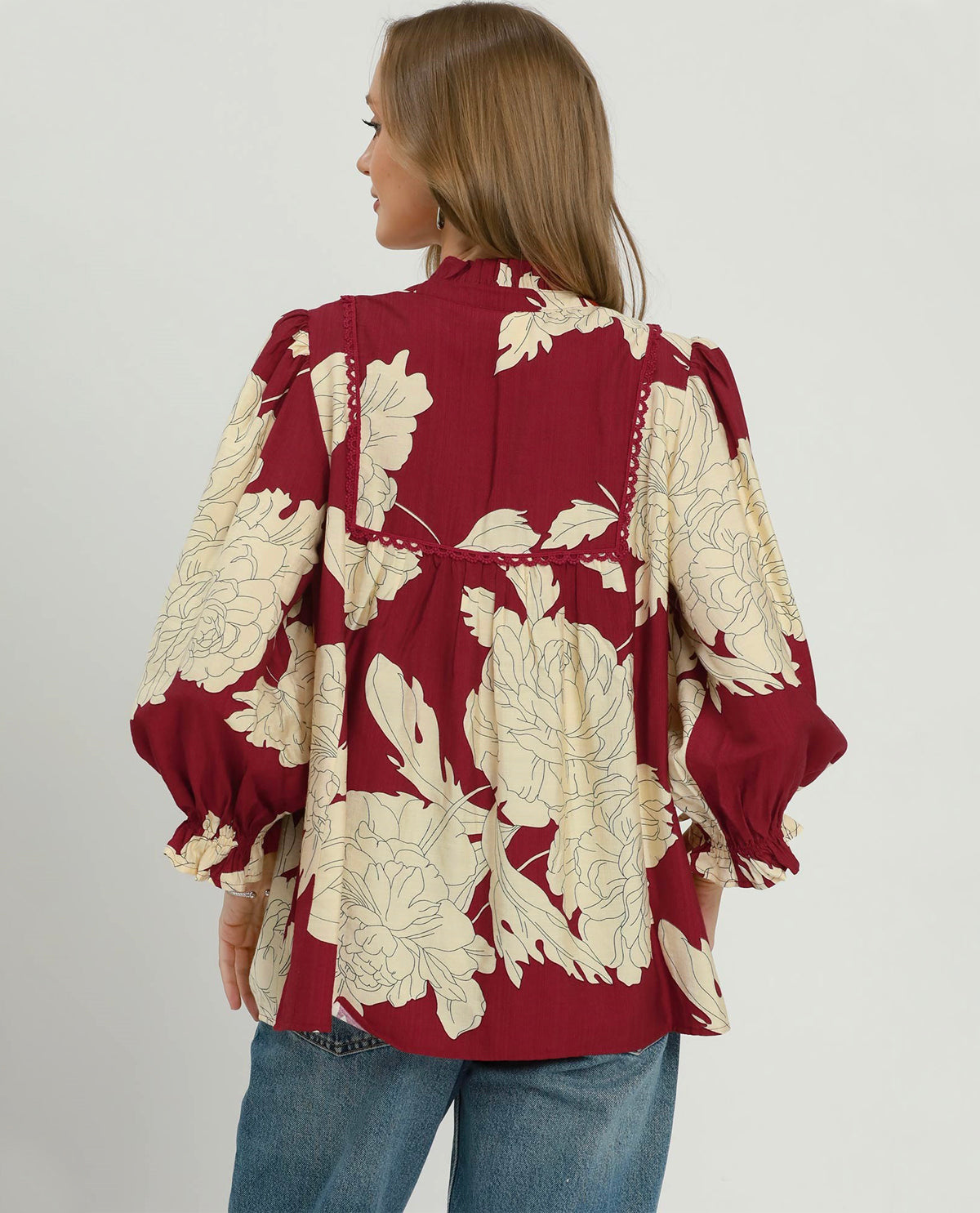 A woman shown from the back wearing a red blouse with a large cream floral print and blue jeans, photographed against a plain light gray studio background.