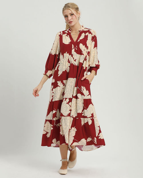 A woman modeling a red midi dress with a beige floral print, featuring a V-neck, 3/4 sleeves, and side pockets, set against a plain light gray studio background.