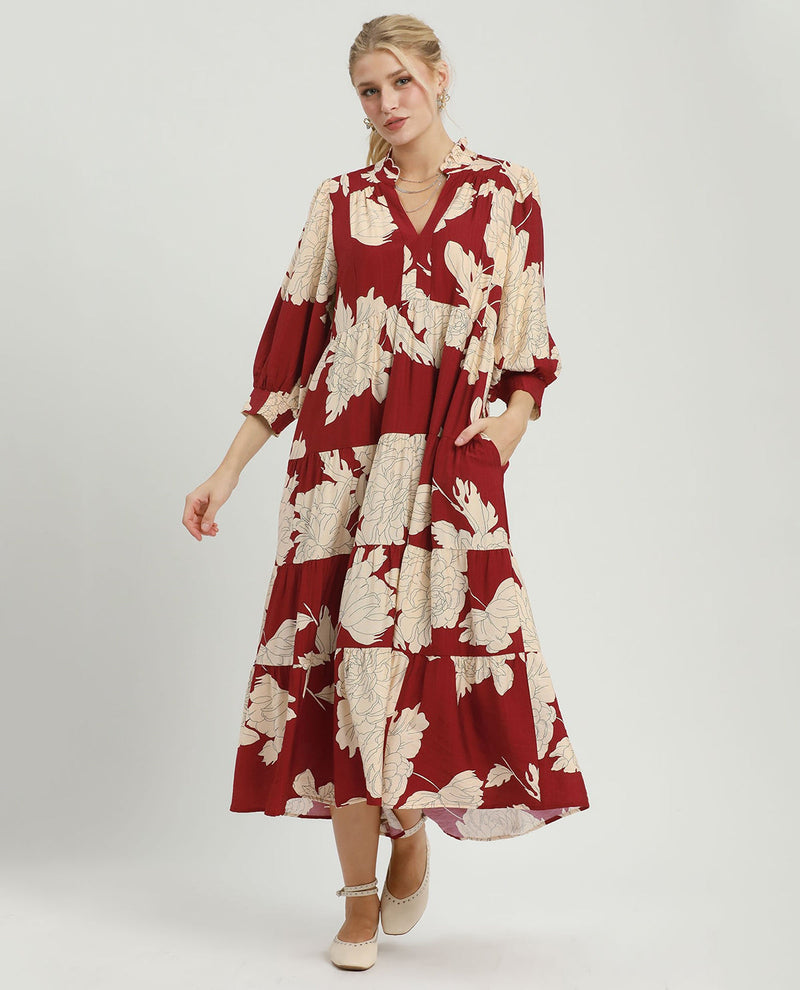 A woman modeling a red midi dress with a beige floral print, featuring a V-neck, 3/4 sleeves, and side pockets, set against a plain light gray studio background.