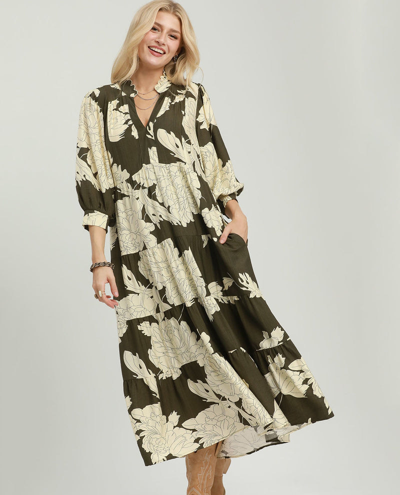 A woman modeling a tiered midi dress with a dark olive green and beige floral pattern, featuring a split neckline, three-quarter sleeves, and side pockets, set against a plain light gray studio background.