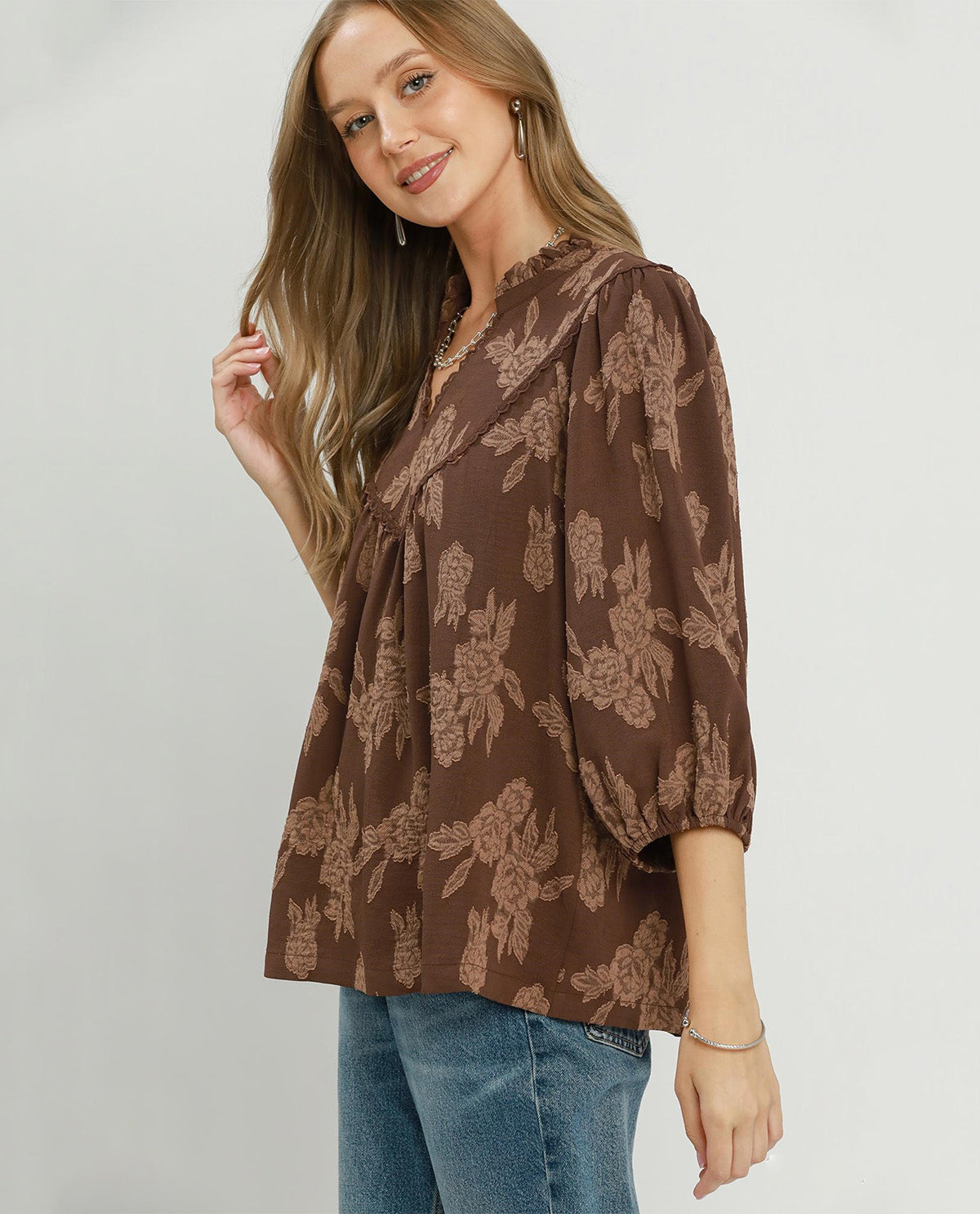 A woman wearing a brown floral print blouse with puff sleeves and blue jeans, standing against a light gray studio background.