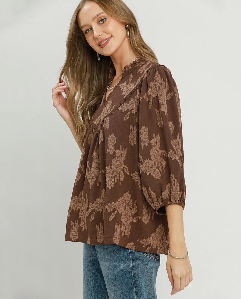 A woman wearing a brown floral print blouse with puff sleeves and blue jeans, standing against a light gray studio background.