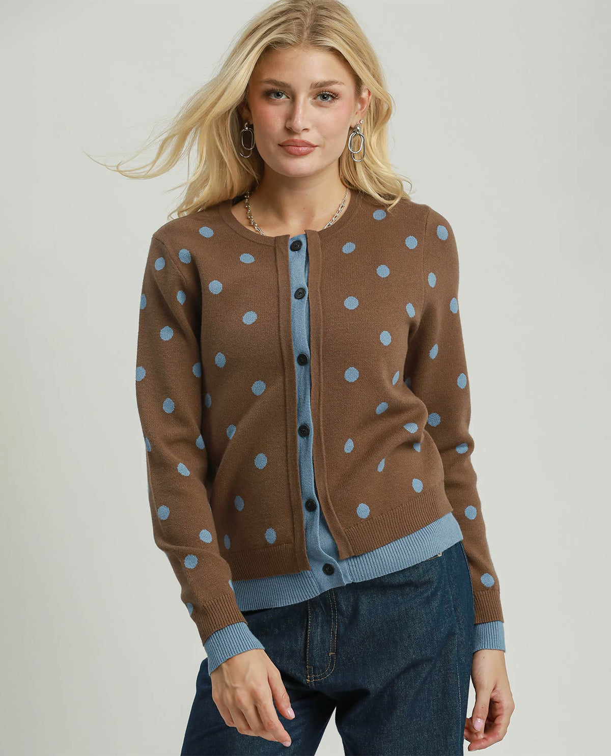 A blonde woman models a brown knit cardigan with light blue polka dots and ribbed trim, worn over a light blue top and dark blue jeans, against a light gray studio background.