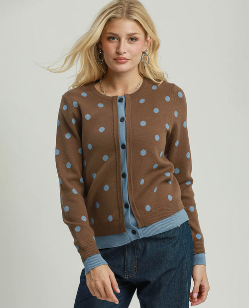 A blonde woman models a brown knit cardigan with light blue polka dots and ribbed trim, worn over a light blue top and dark blue jeans, against a light gray studio background.