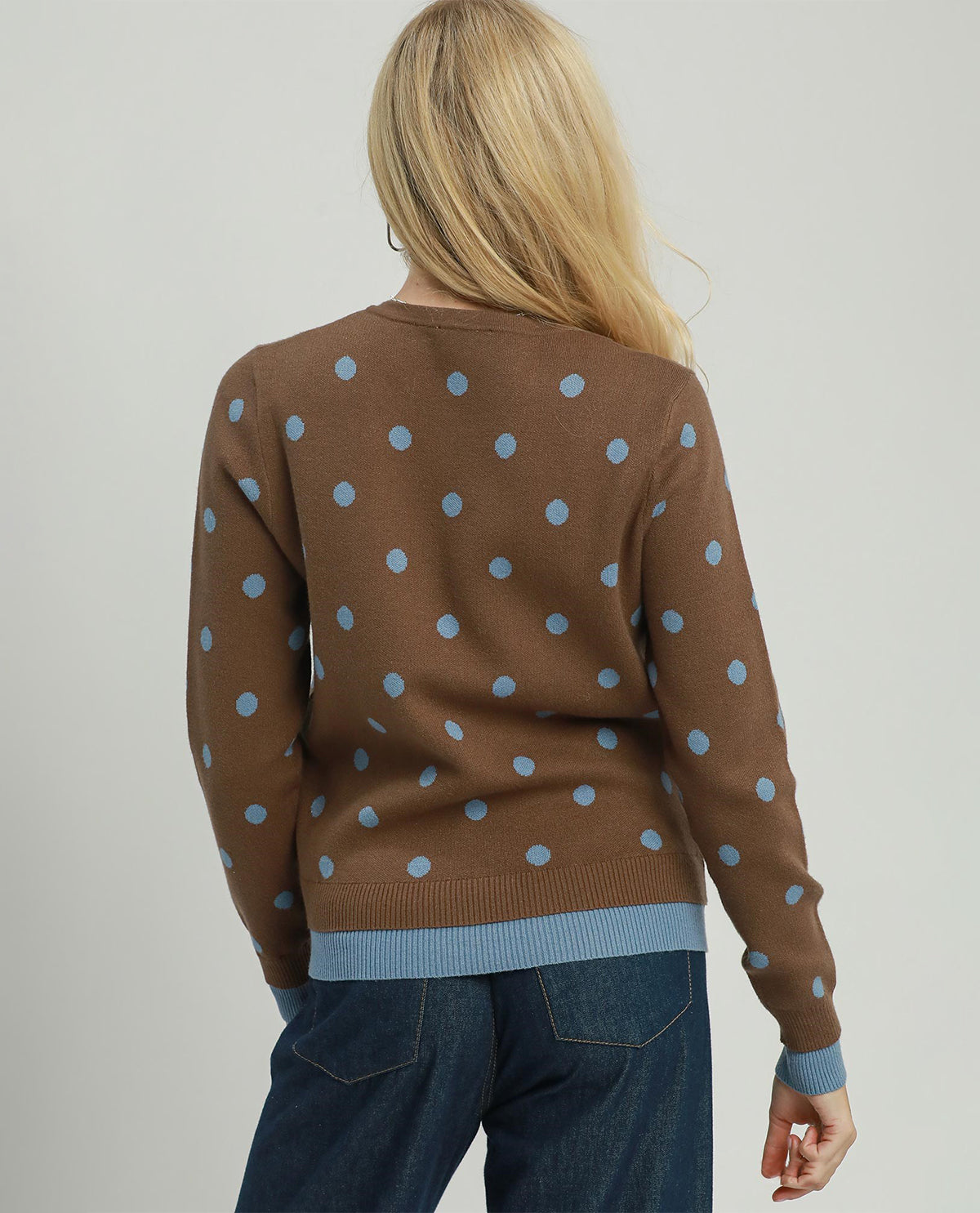 A woman viewed from behind wearing a brown knit sweater with light blue polka dots and ribbed trim, paired with dark blue jeans, against a plain light gray studio background.