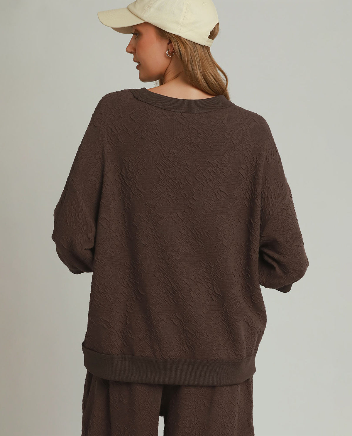 A woman shown from the back wearing a textured dark brown crewneck sweatshirt and matching brown pants, paired with a cream-colored baseball cap, against a plain light gray studio background.