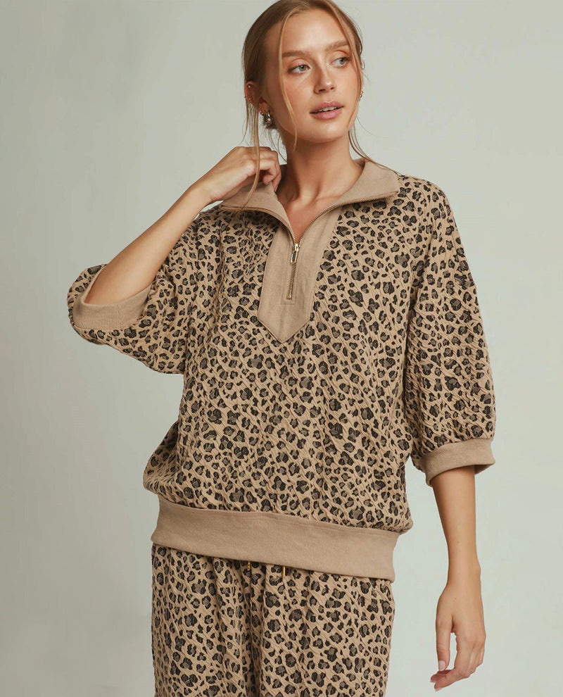 A woman modeling a matching beige and brown leopard-print lounge set, featuring a half-zip pullover top and matching pants, against a solid light gray background.
