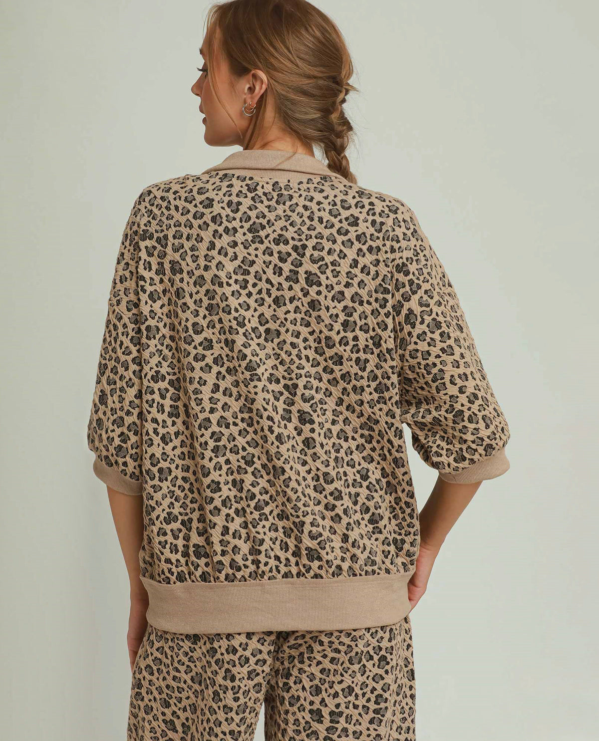 A woman shown from the back wearing a beige and brown leopard-print lounge set with a collared top and matching pants, against a plain light gray studio background.