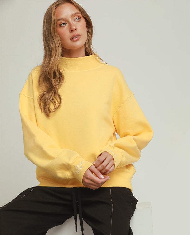 A woman wearing a pale yellow mock-neck sweatshirt and black drawstring pants, seated against a light gray studio background.