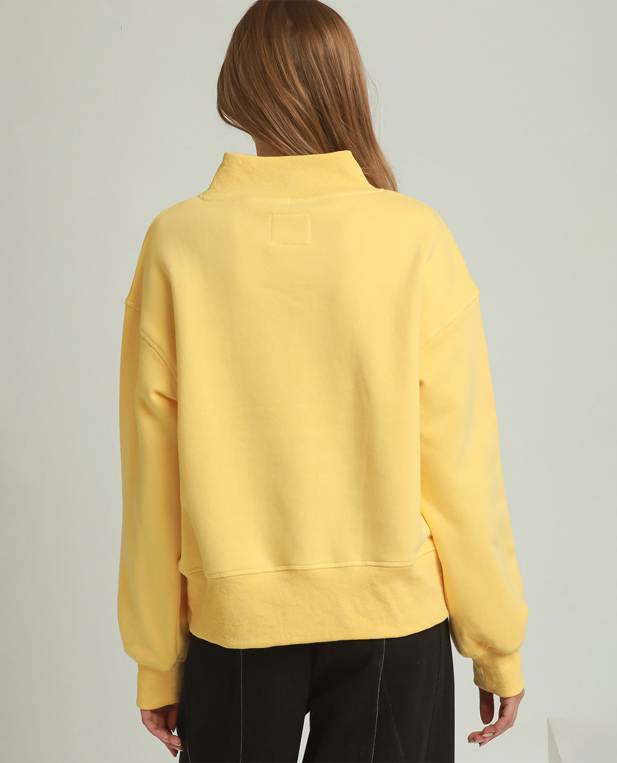 A woman viewed from the back wearing a pale yellow mock-neck sweatshirt and black pants, set against a plain light gray studio background.