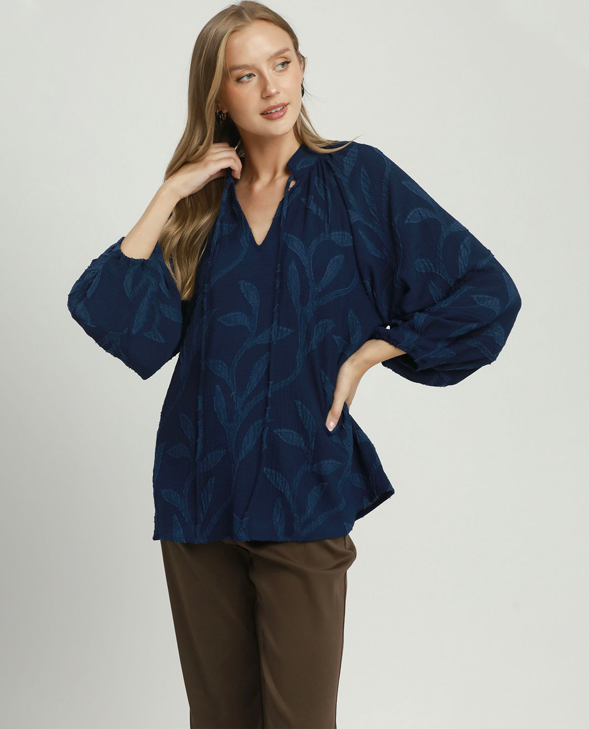 A woman modeling a navy blue blouse with a tonal leaf pattern, featuring voluminous elbow-length sleeves and a gathered V-neckline, paired with brown trousers against a light gray studio background.