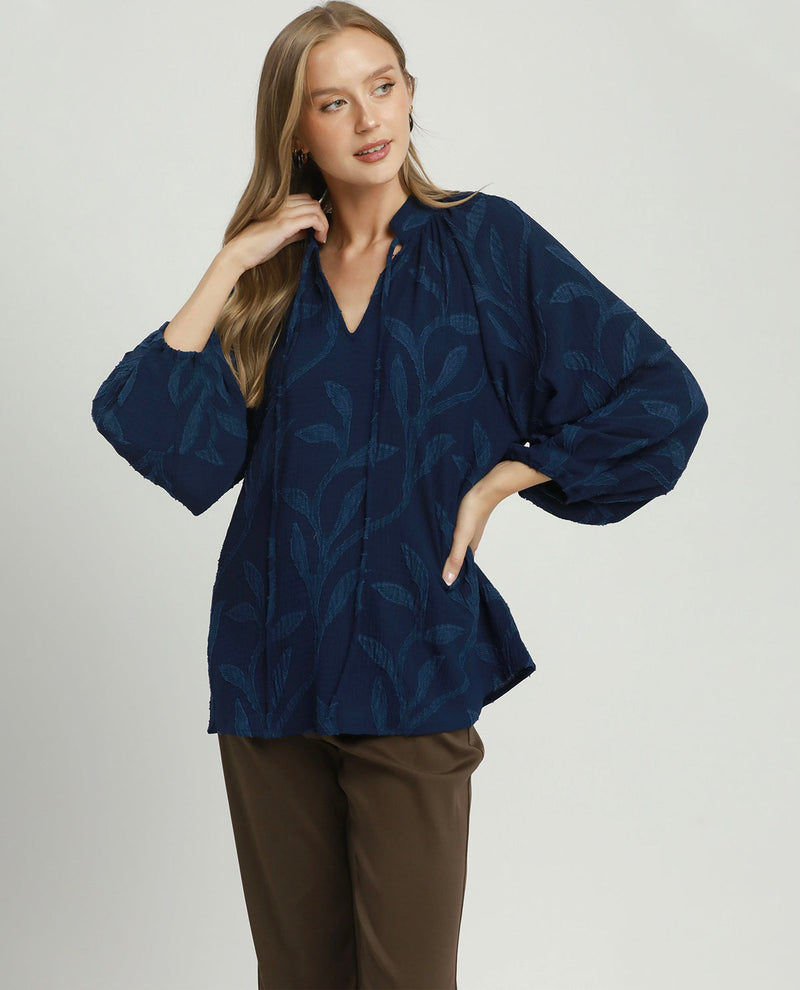 A woman modeling a navy blue blouse with a tonal leaf pattern, featuring voluminous elbow-length sleeves and a gathered V-neckline, paired with brown trousers against a light gray studio background.