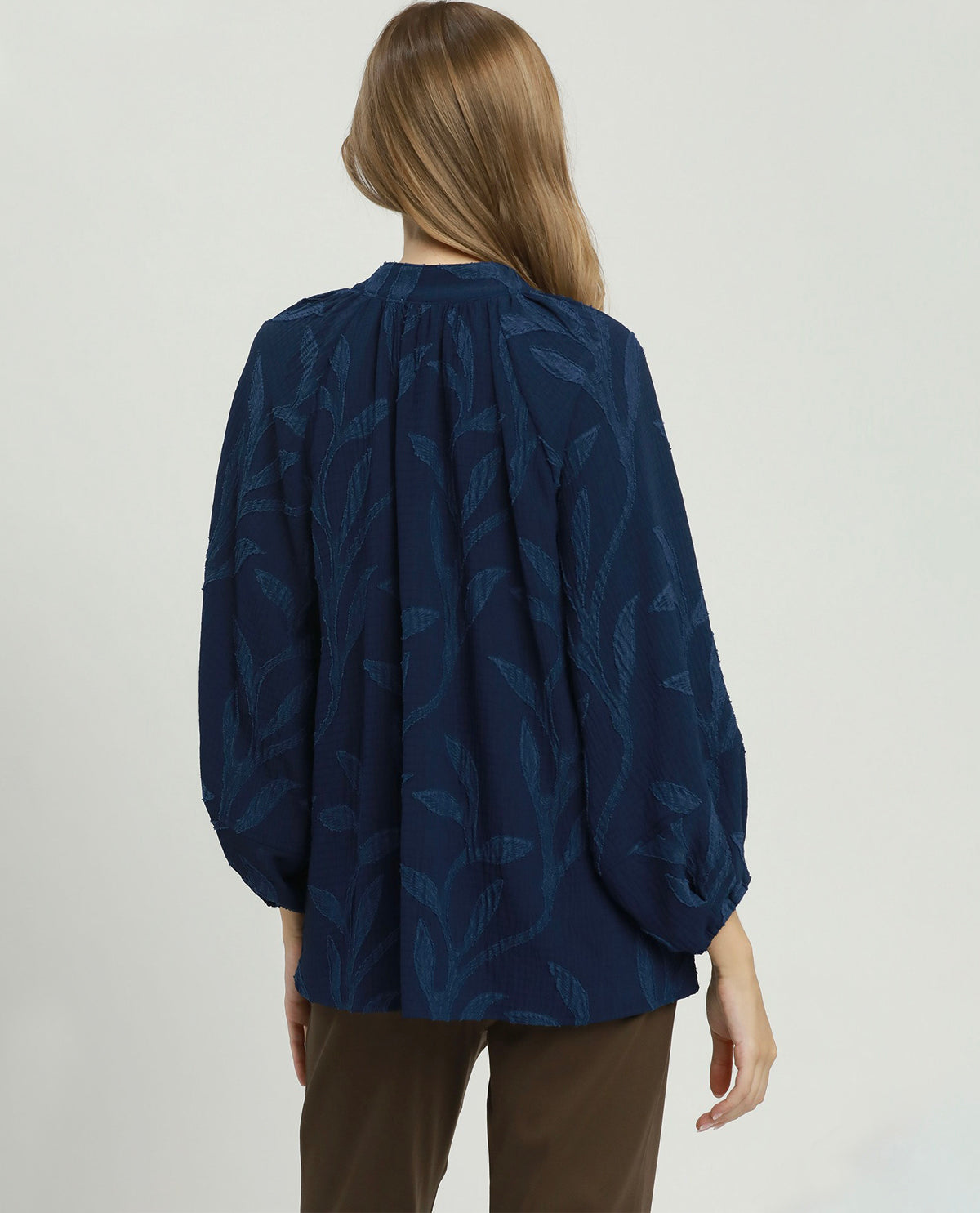 A woman viewed from the back wearing a navy blue long-sleeve blouse with a tonal leaf pattern and brown trousers, set against a plain light gray studio background.