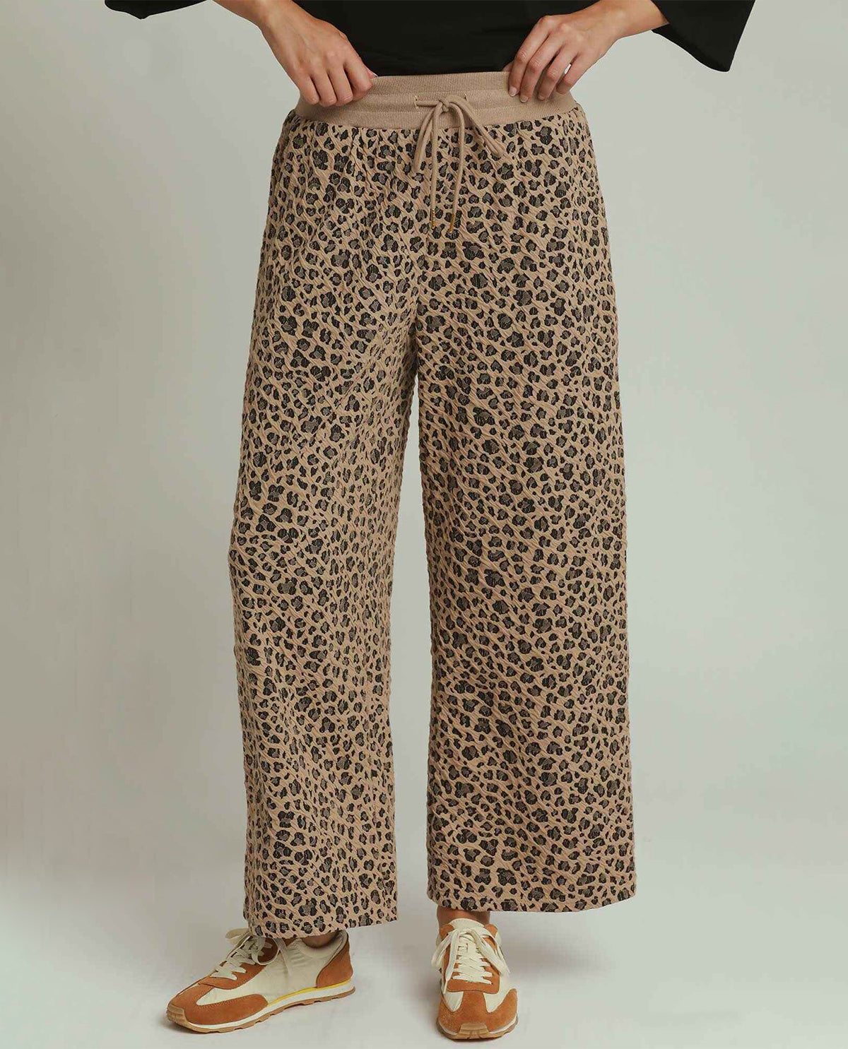 A person wearing wide-leg leopard-print pants with a drawstring waistband, paired with brown and cream sneakers, shown from the waist down against a plain light gray studio background.